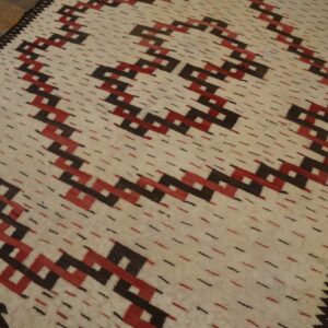 Cream rug with interlocking red and dark brown geometric patterns, accented by dashes, on wood plank flooring.