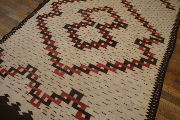 Cream rug with interlocking red and dark brown geometric patterns, accented by dashes, on wood plank flooring.
