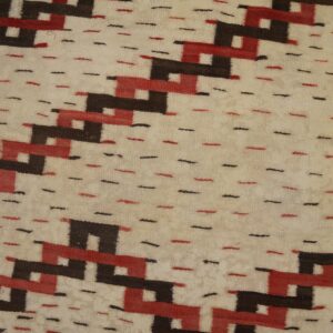 Cream flatweave rug with repeating red and dark brown geometric patterns and a sawtooth edge on wood planks.