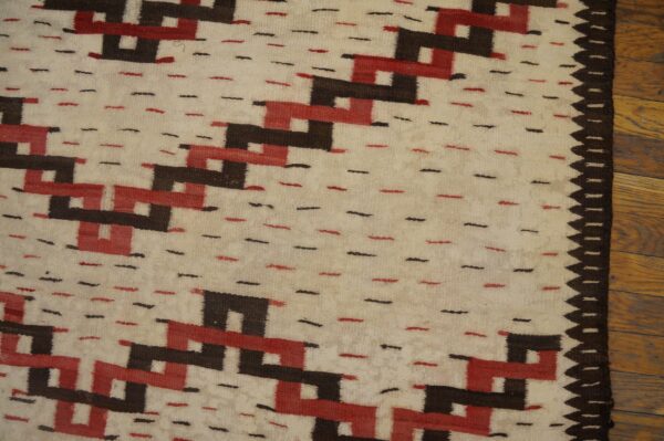 Cream flatweave rug with repeating red and dark brown geometric patterns and a sawtooth edge on wood planks.