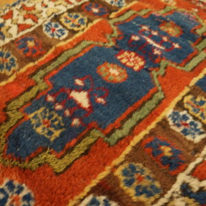 Close-up of a high-pile rug featuring blue, rust, brown, and green geometric motifs on wooden floorboards.