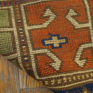 A folded geometric rug in rust, green, and cream colors rests on distressed light wood flooring.
