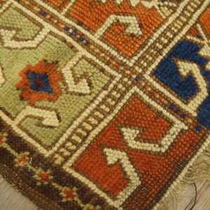 Geometric low-pile rug corner in orange, sage green, and blue resting on distressed light wooden flooring.