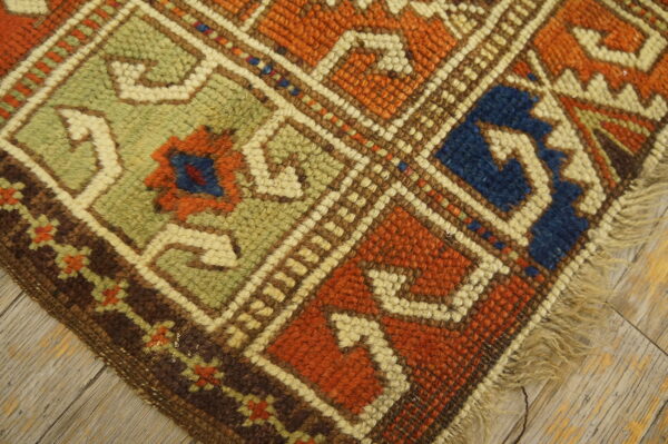 Geometric low-pile rug corner in orange, sage green, and blue resting on distressed light wooden flooring.