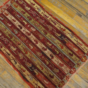 Low-pile rug featuring complex red, green, and tan vertical stripes on faded wood flooring.