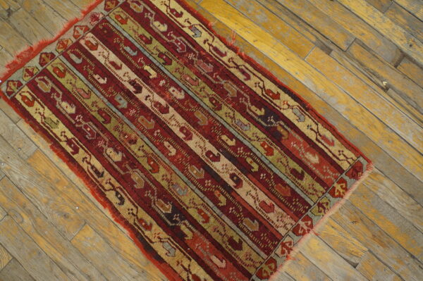 Low-pile rug featuring complex red, green, and tan vertical stripes on faded wood flooring.