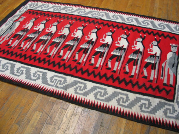 Red, black, gray, and white flatweave rug featuring stylized figures and a meander border on a wood floor.