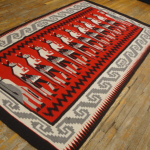 Red flatweave rug with repeating stylized figures and geometric borders rests on worn hardwood flooring.