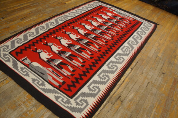 Red flatweave rug with repeating stylized figures and geometric borders rests on worn hardwood flooring.