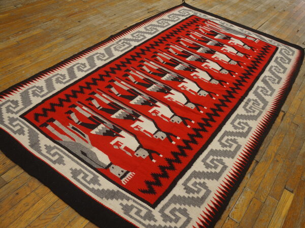 Red flatweave rug with repeating black and white figures and a grey key pattern border on wood planks.