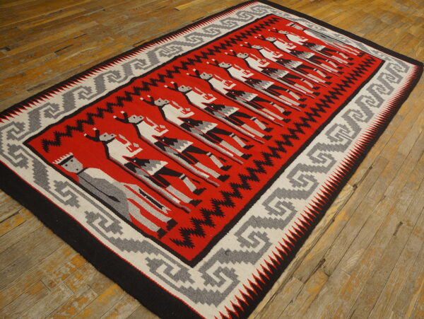 Red, gray, and black flatweave rug featuring stylized figures and a geometric border on distressed wood flooring.
