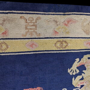 Detail of a blue rug showing stylized motifs and a patterned border in cream, yellow, pink, and tan.