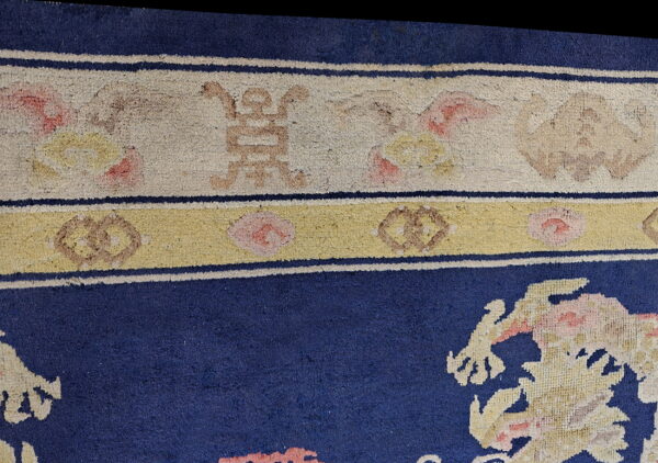 Detail of a blue rug showing stylized motifs and a patterned border in cream, yellow, pink, and tan.