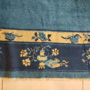 Plush teal rug with ornate navy and gold floral borders, visible fringe, resting on light tile.