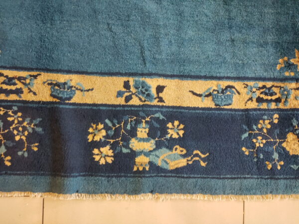 Plush teal rug with ornate navy and gold floral borders, visible fringe, resting on light tile.