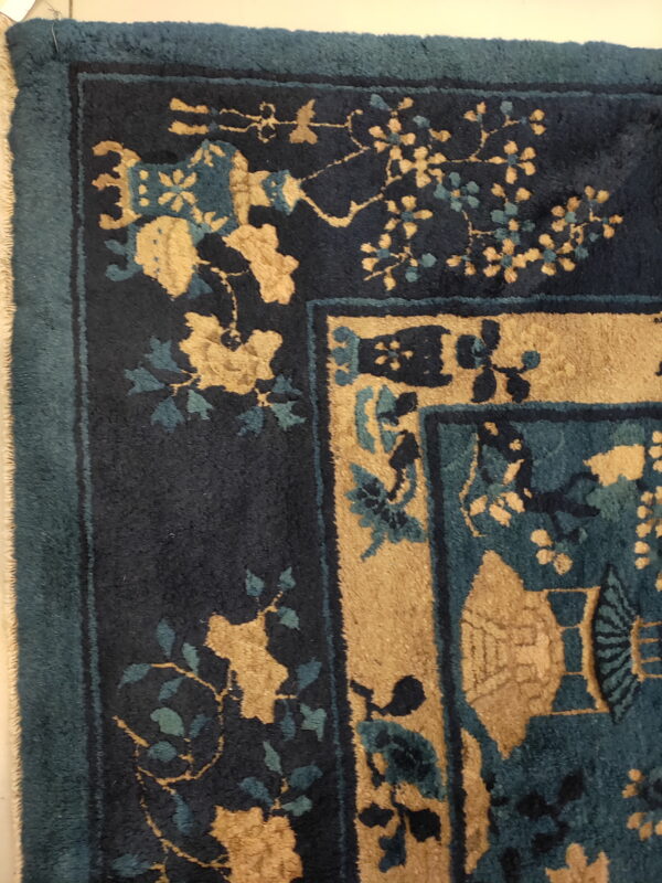 Detail of a plush rug showing dark blue, teal, and gold floral corner design and fringed edge.