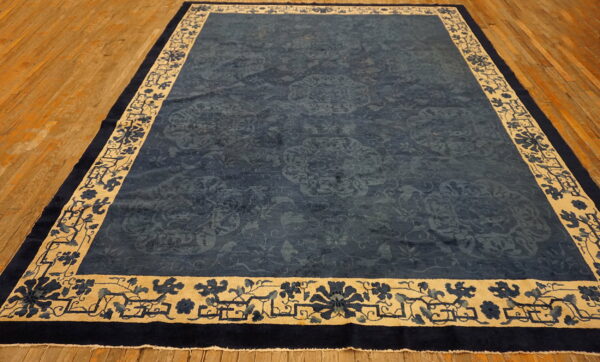 Navy blue rug featuring subtle medallions and a wide cream border with dark floral patterns rests on wood planks.