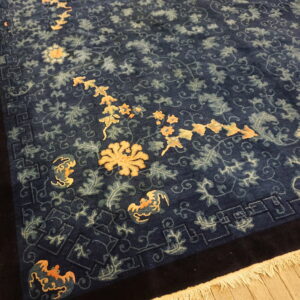 Dark blue rug with stylized gold and sage floral design and key border rests on light wood planks.