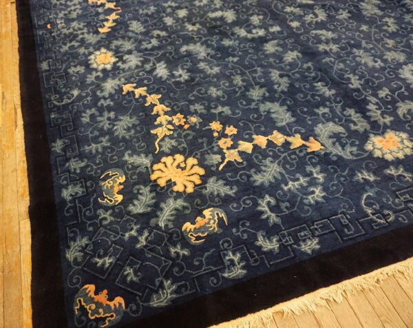 Dark blue rug with stylized gold and sage floral design and key border rests on light wood planks.