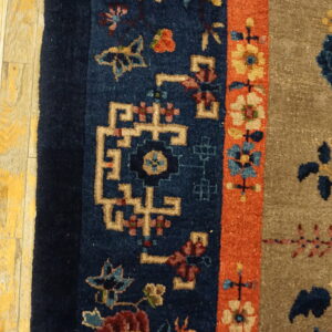 Textured, detailed rug featuring tan, blue, and orange floral borders next to worn yellow wood flooring.