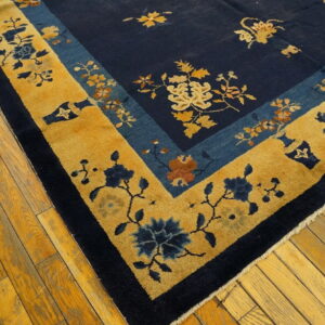 Close-up of a navy and gold floral rug with wide borders lying diagonally on yellow plank flooring.