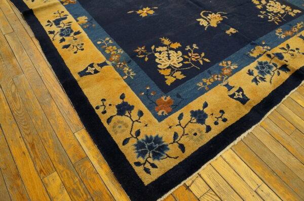 Close-up of a navy and gold floral rug with wide borders lying diagonally on yellow plank flooring.