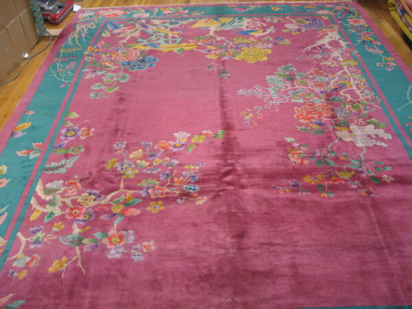 Deep pink rug with a wide teal border featuring multi-colored floral and bird motifs on wood flooring.
