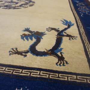 Angled view of a rug with a cream field, blue dragons, and a geometric fret pattern border.