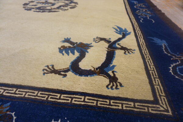 Angled view of a rug with a cream field, blue dragons, and a geometric fret pattern border.