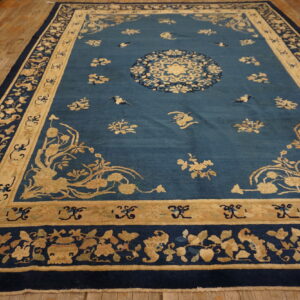 Rectangular blue rug with extensive tan and navy floral borders on a distressed wood floor.