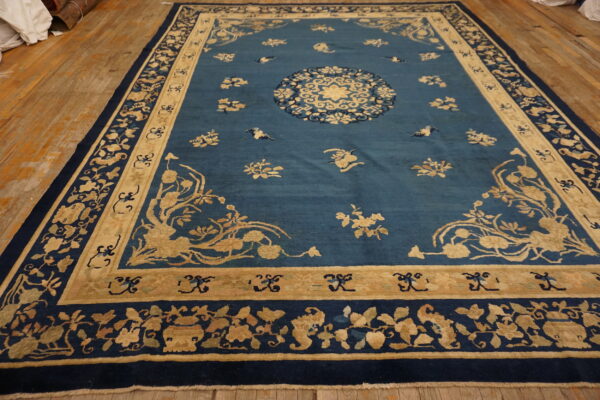 Rectangular blue rug with extensive tan and navy floral borders on a distressed wood floor.