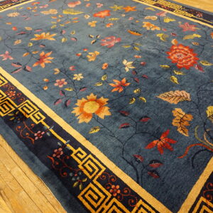 Teal-blue rug with detailed floral vines and gold geometric borders lies on light hardwood flooring.