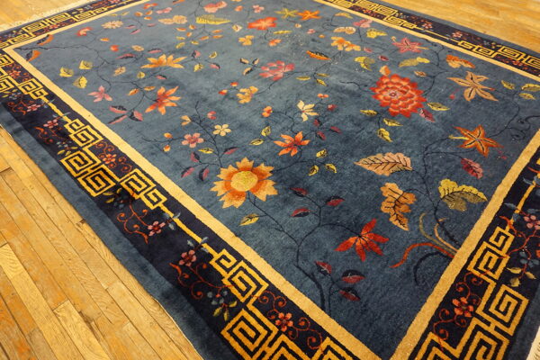 Teal-blue rug with detailed floral vines and gold geometric borders lies on light hardwood flooring.