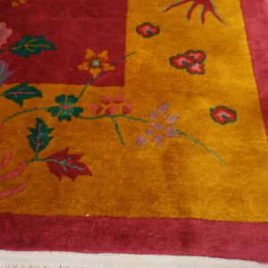 Corner detail of a plush red and gold floral rug with green accents resting on light wood flooring.