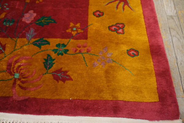 Corner detail of a plush red and gold floral rug with green accents resting on light wood flooring.