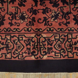 A close-up of a high-pile rug border in rust, black, and beige, over light wooden flooring.
