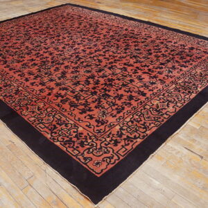 Red-orange rug with black scrollwork and a wide black border resting on distressed light wooden floors.