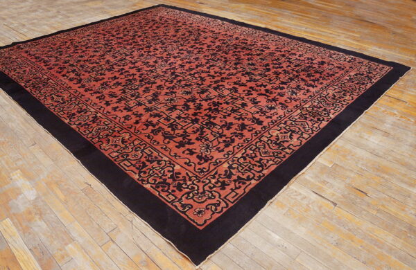 Red-orange rug with black scrollwork and a wide black border resting on distressed light wooden floors.