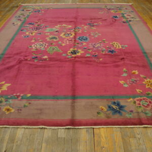 Pink rug with multicolored floral design and taupe border lying flat on distressed wood floor.