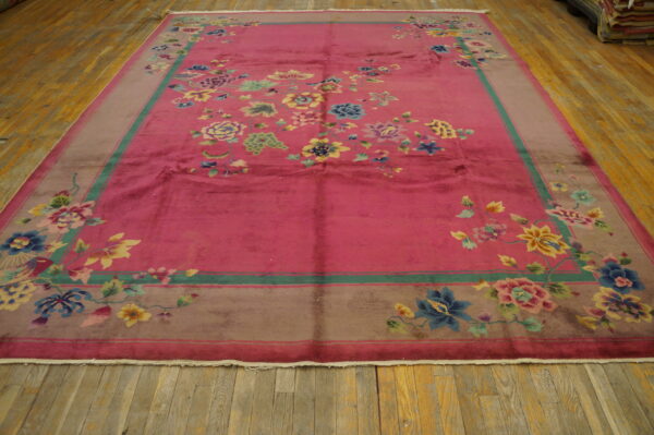 Pink rug with multicolored floral design and taupe border lying flat on distressed wood floor.