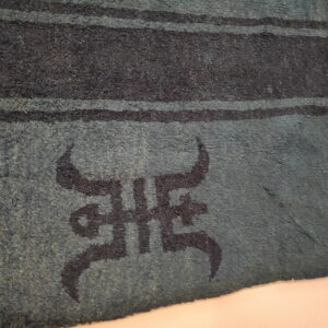Textured teal rug displayed at an angle, showing dark horizontal stripes and a central dark symbolic motif.