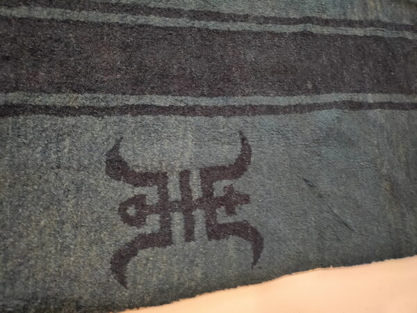 Textured teal rug displayed at an angle, showing dark horizontal stripes and a central dark symbolic motif.