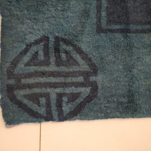 Textured teal rug with a dark blue geometric pattern detail resting on light tiled flooring.