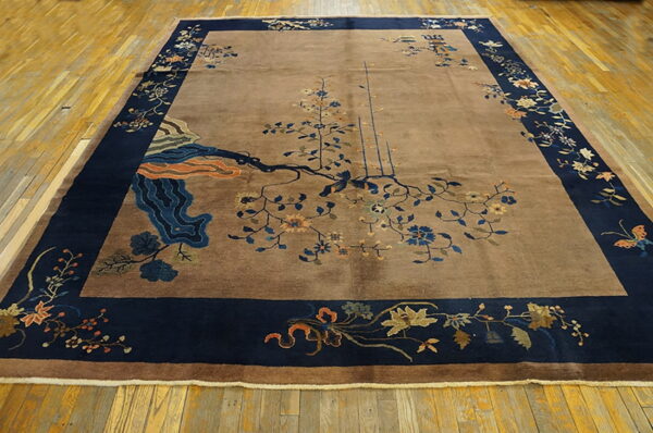A plush tan rug with a wide navy blue floral border lies on light wooden floor planks.
