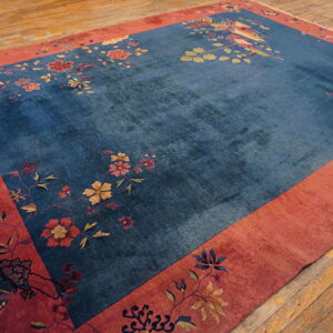 A deep blue rug with multicolored floral patterns and a bird design, framed by a wide red border on wood floors.