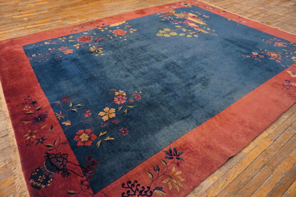A deep blue rug with multicolored floral patterns and a bird design, framed by a wide red border on wood floors.
