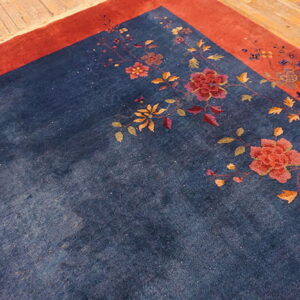Corner view of a navy rug featuring a winding botanical design and a wide rusty red border on wood flooring.