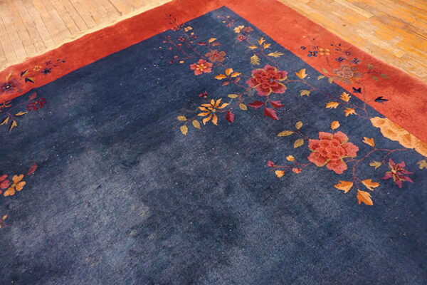 Corner view of a navy rug featuring a winding botanical design and a wide rusty red border on wood flooring.