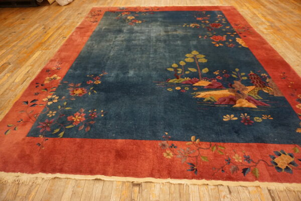An ornate blue rug featuring a scenic motif and a wide red floral border on wood plank flooring.