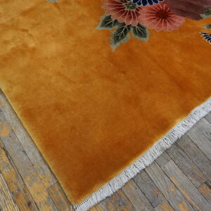 Plush golden-orange rug with pink and blue floral detail on distressed gray wood floor.